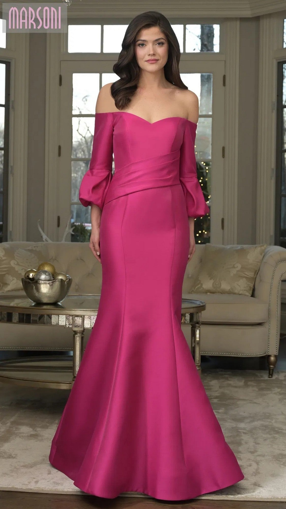 A woman poses indoors in the Marsoni MV1355 long formal dress in fuchsia, featuring an off-the-shoulder neckline, three-quarter puff sleeves, and a subtle mermaid silhouette, exuding elegance.
